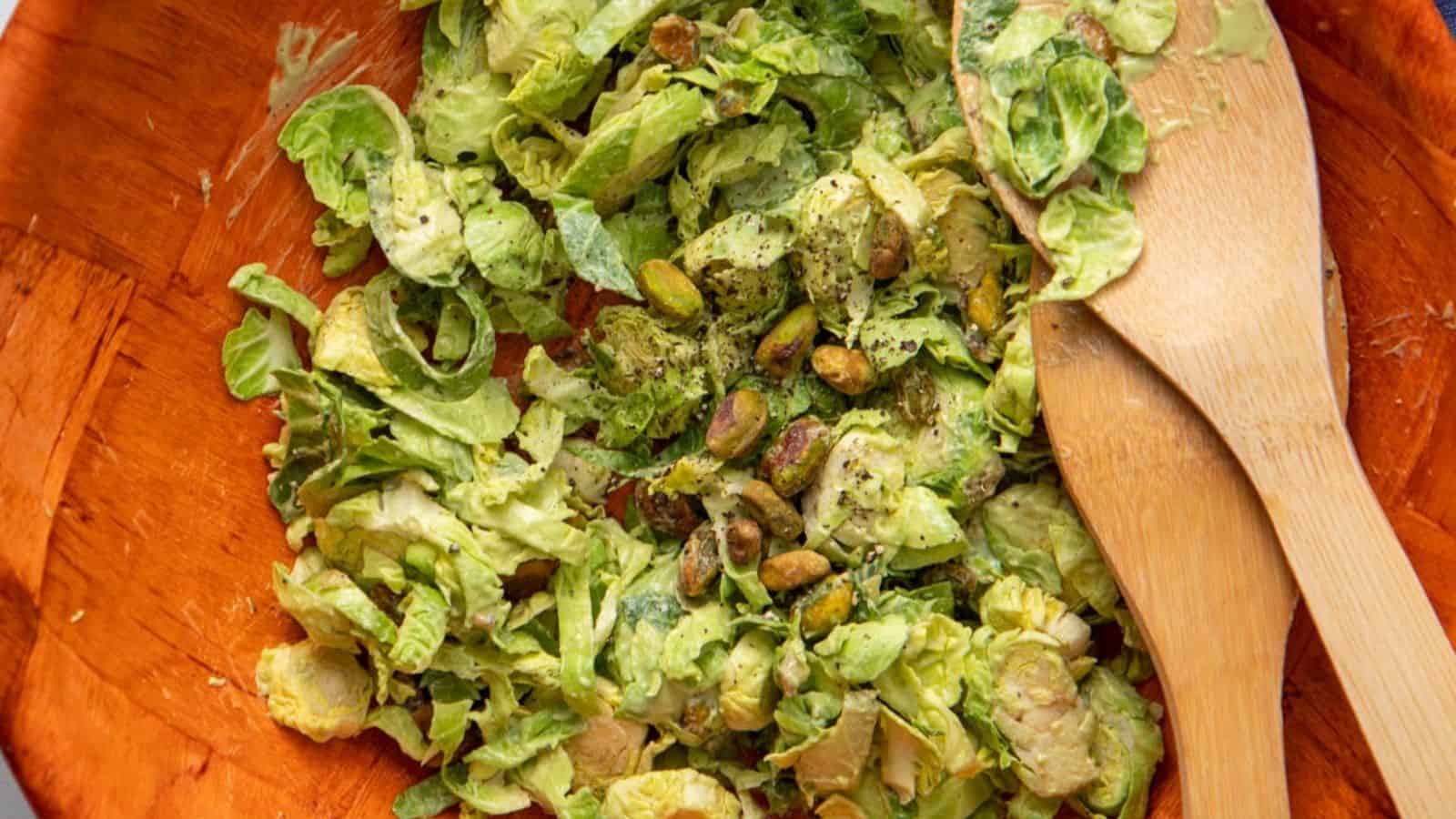 Chopped Brussels sprouts salad with pistachios, lightly dressed, in a wooden bowl with wooden serving utensils.
