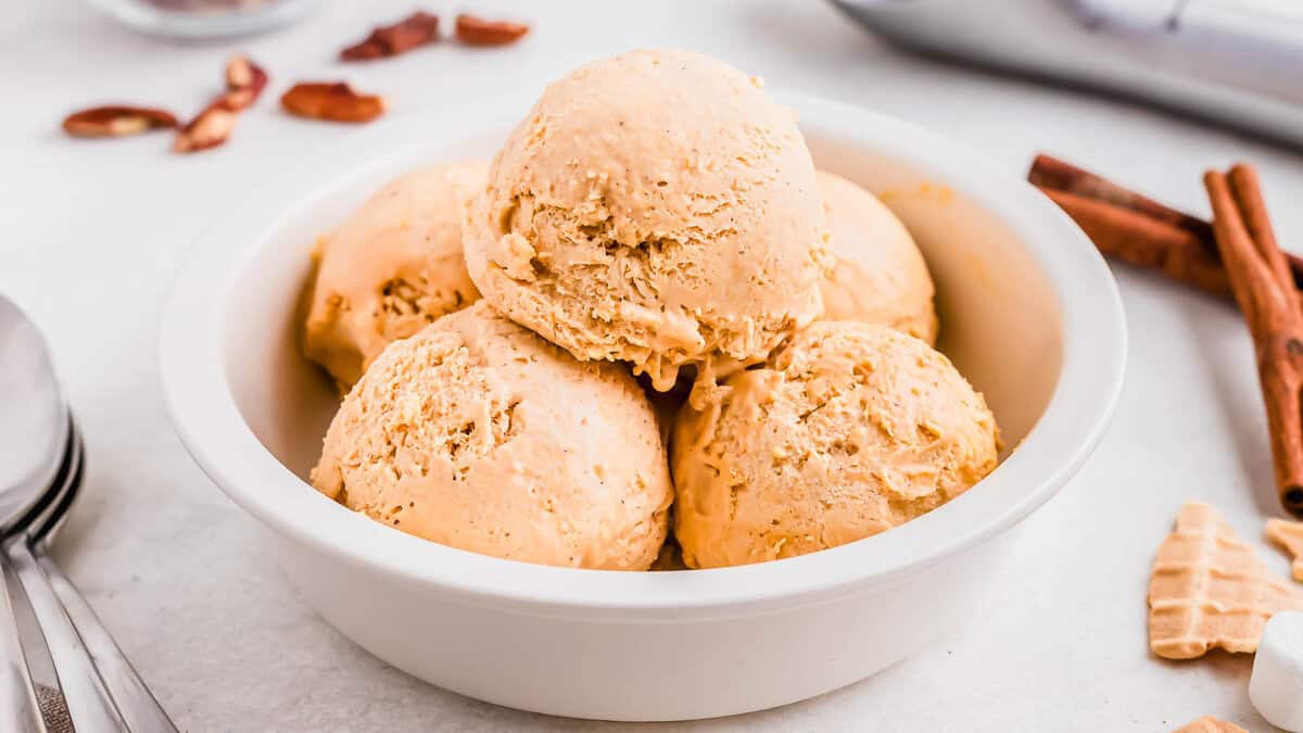 A white bowl filled with five scoops of light orange ice cream, with cinnamon sticks and pecan pieces in the background.