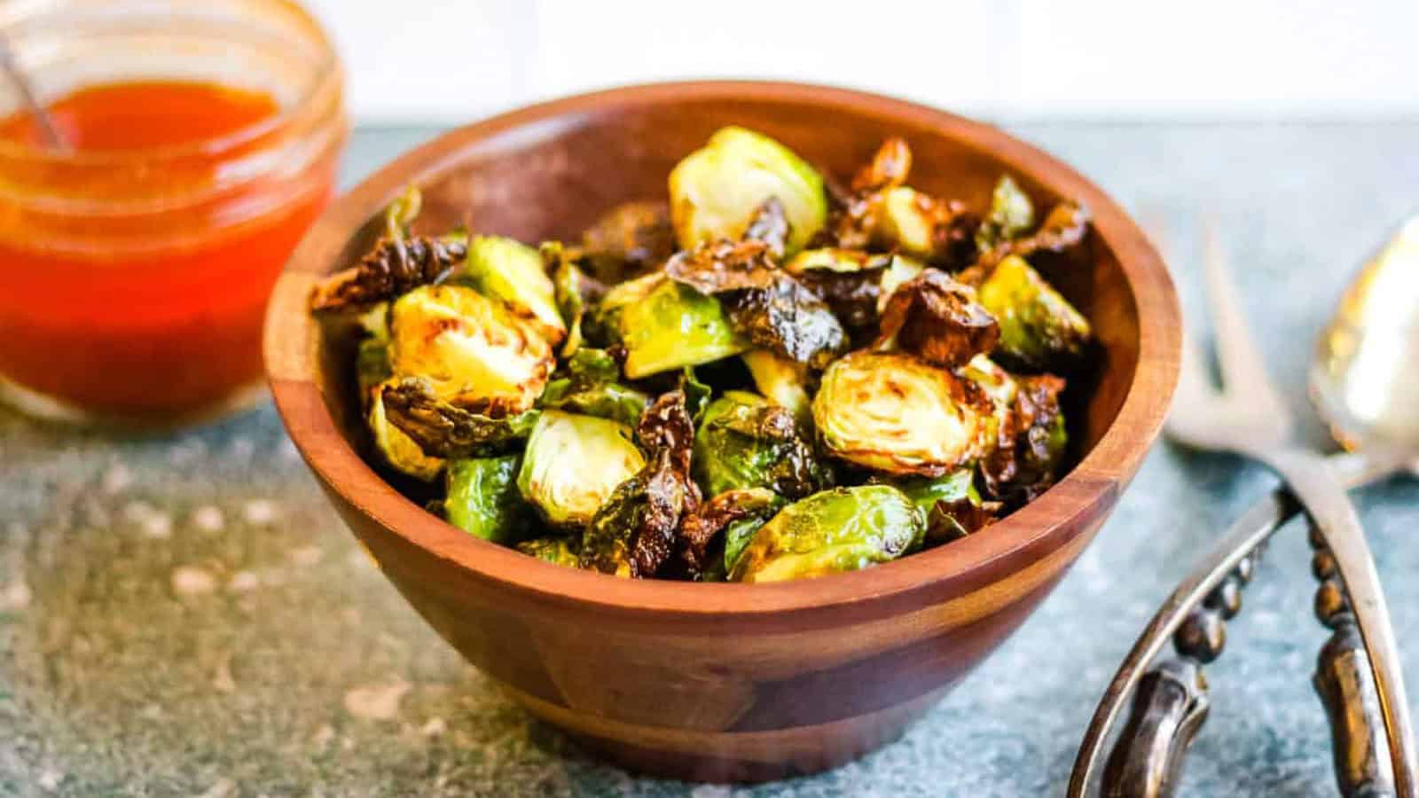 A wooden bowl filled with roasted Brussels sprouts sits on a countertop next to a jar of orange sauce and a pair of metal tongs.