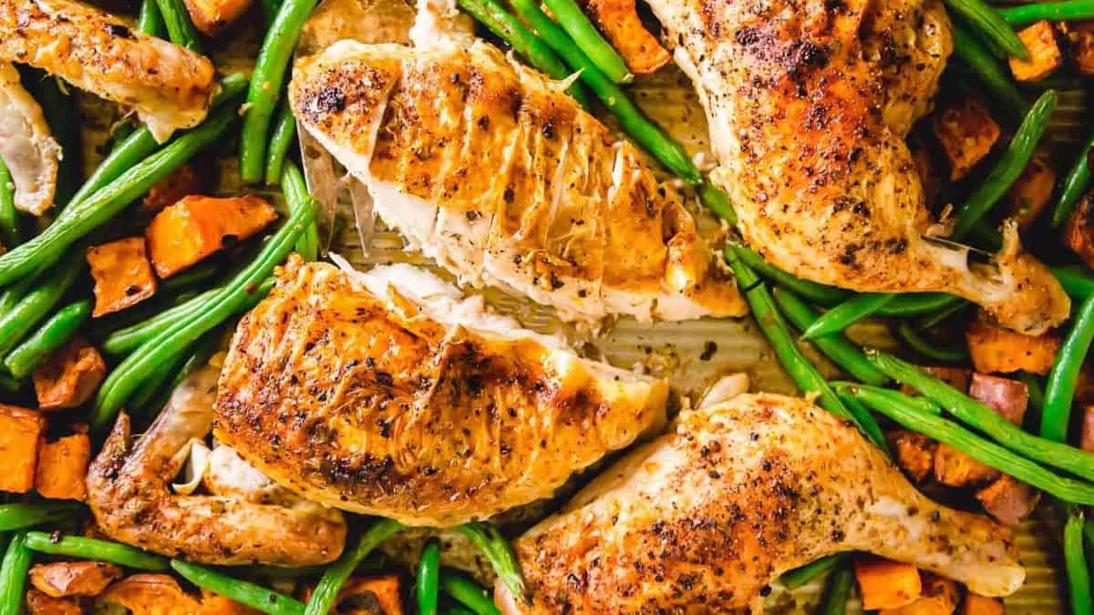 Roast chicken pieces with crispy skin, surrounded by green beans and roasted sweet potato cubes on a baking sheet.