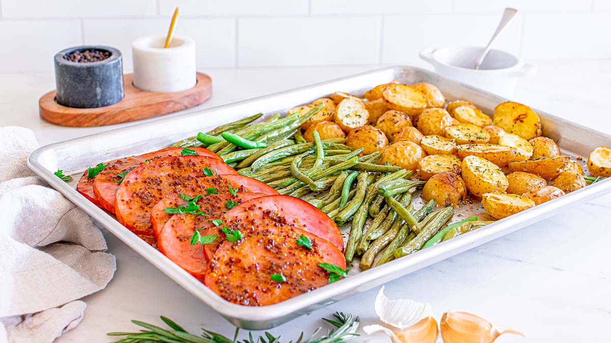 Sheet pan with sliced ham, roasted green beans, and seasoned potato halves, garnished with herbs, on a white kitchen counter.