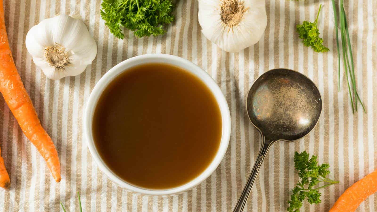 A bowl of brown broth sits on a striped cloth, surrounded by carrots, garlic bulbs, parsley, and a metal ladle.