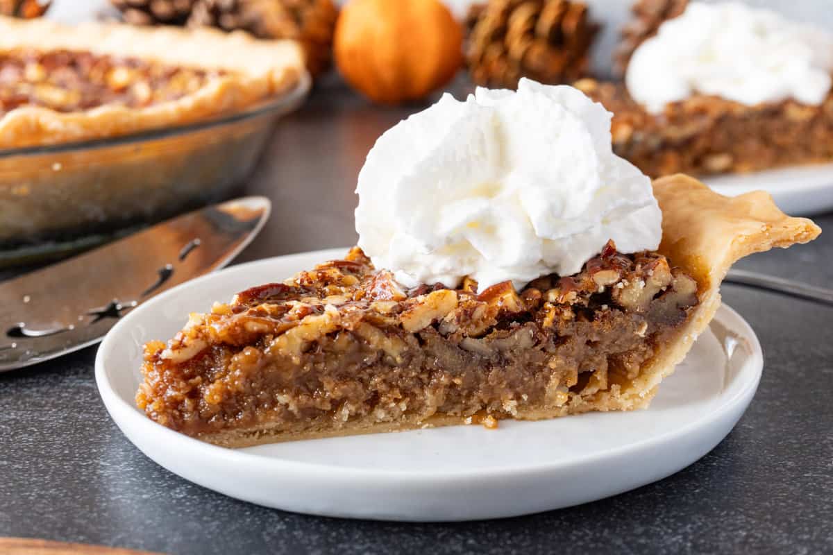 A slice of pecan pie topped with whipped cream on a white plate, with a pie server and pie dish in the background.