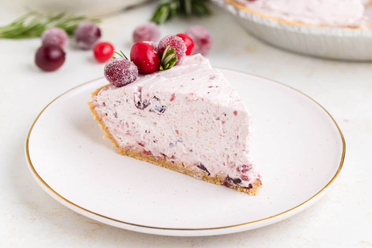 A slice of cranberry cream pie on a white plate, topped with sugared cranberries, with more cranberries in the background.