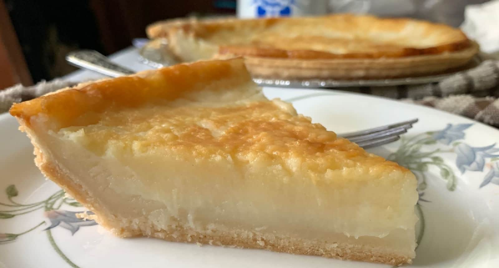 A slice of custard pie on a decorative plate with the rest of the pie in the background.