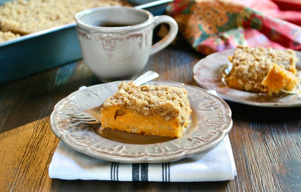 A slice of crumb-topped coffee cake on a plate with a fork, next to a cup and another plate, set on a wooden table.