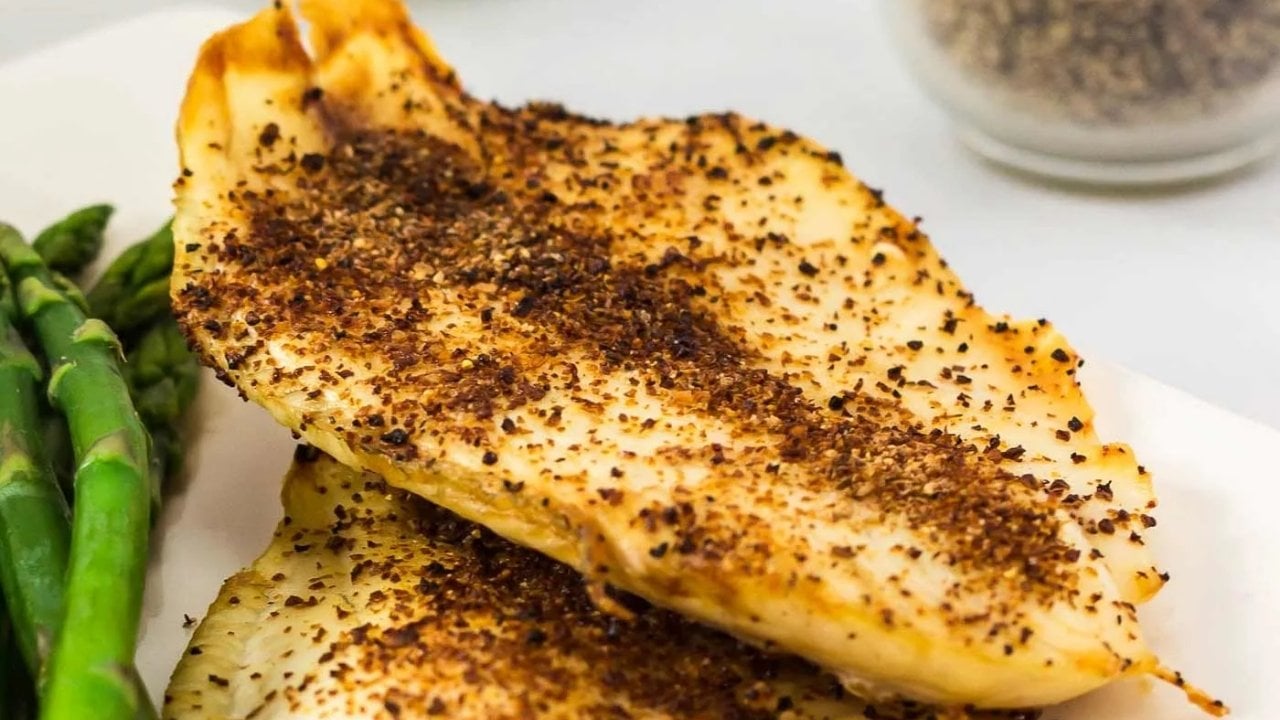 Two seasoned, cooked chicken breasts are served on a white plate with a side of asparagus spears. A container of pepper is visible in the background.