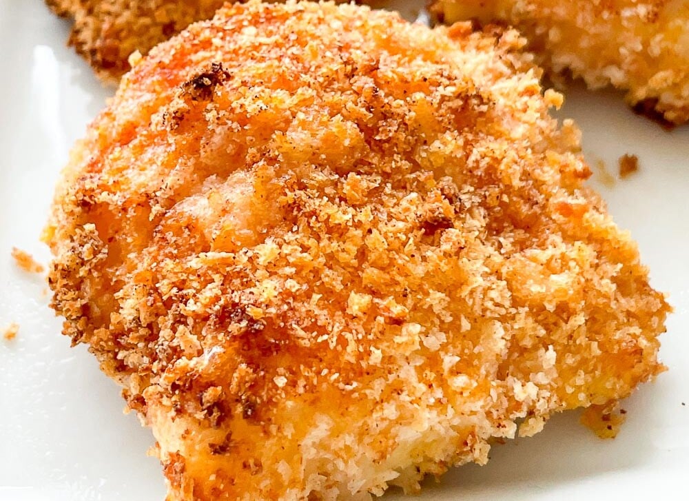 A close-up of a breaded and baked chicken thigh on a white plate, featuring a golden brown, crispy coating.
