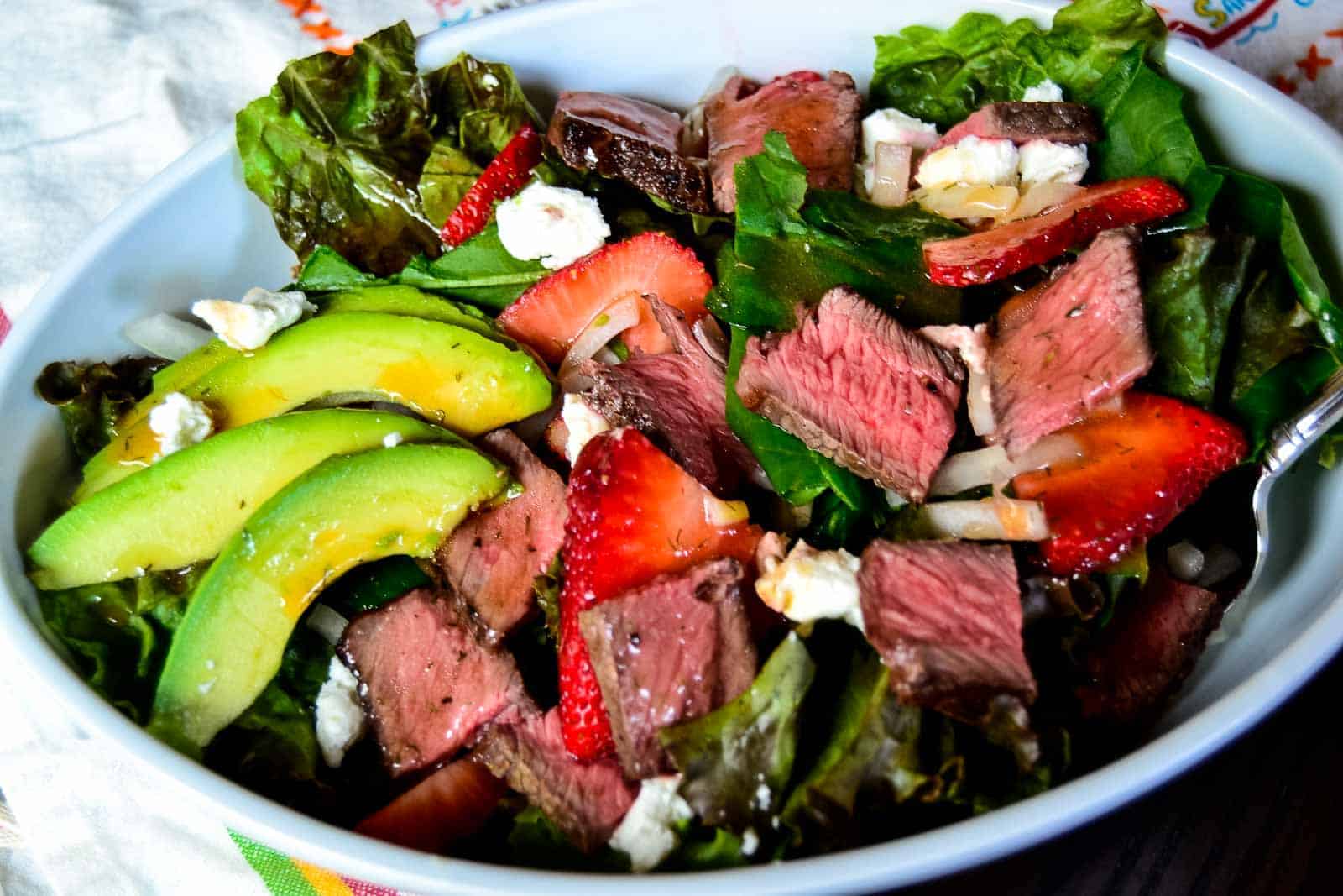A salad bowl with sliced steak, avocado, strawberries, feta cheese, and leafy greens.