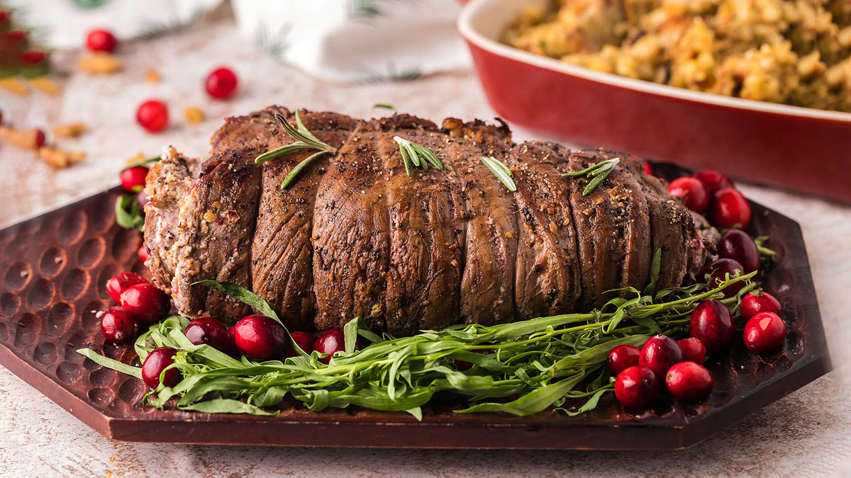 A cooked beef roast garnished with rosemary, fresh greens, and cranberries on a brown platter, with stuffing in the background.