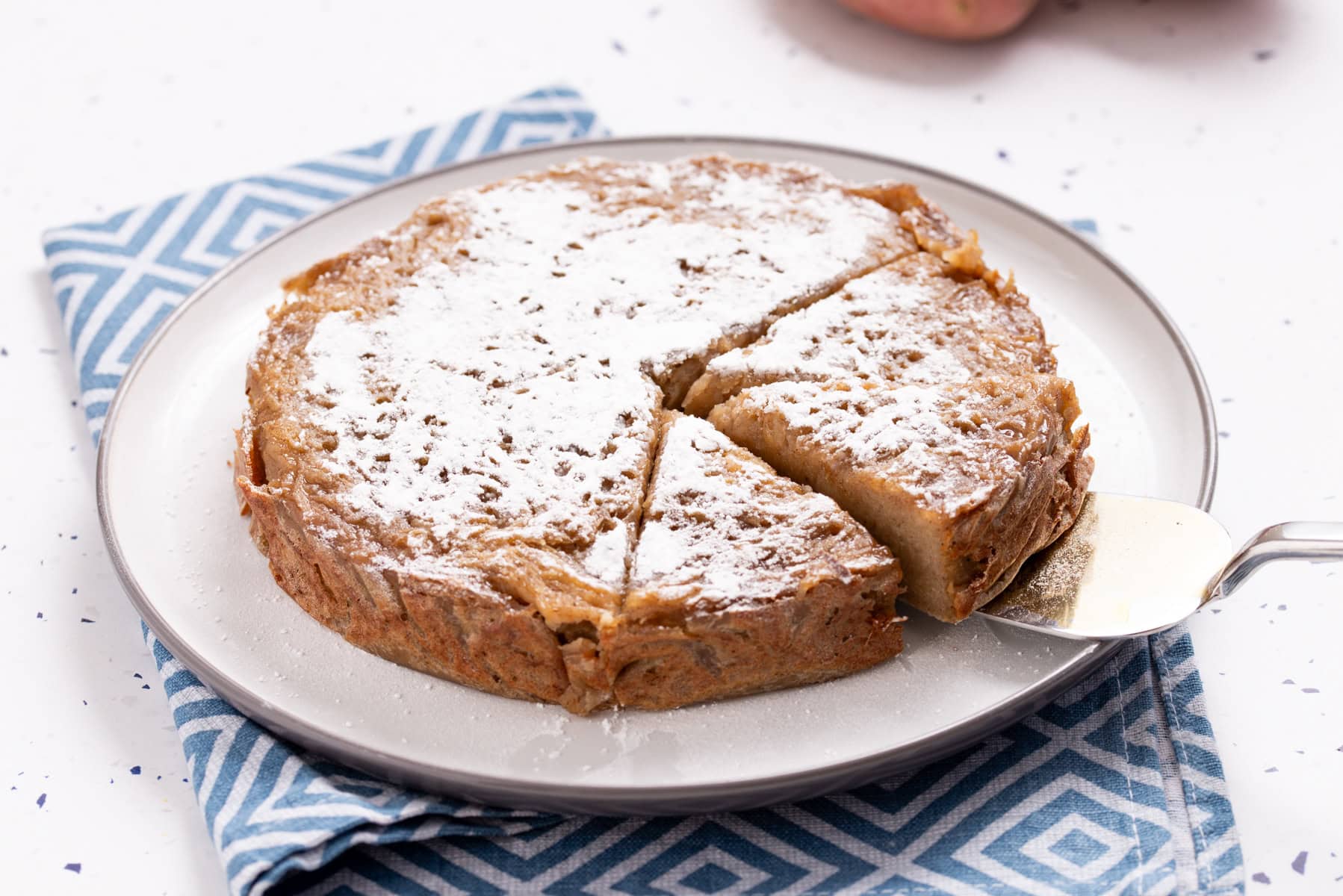 A round cake dusted with powdered sugar, partially sliced, sits on a plate atop a blue-patterned napkin.