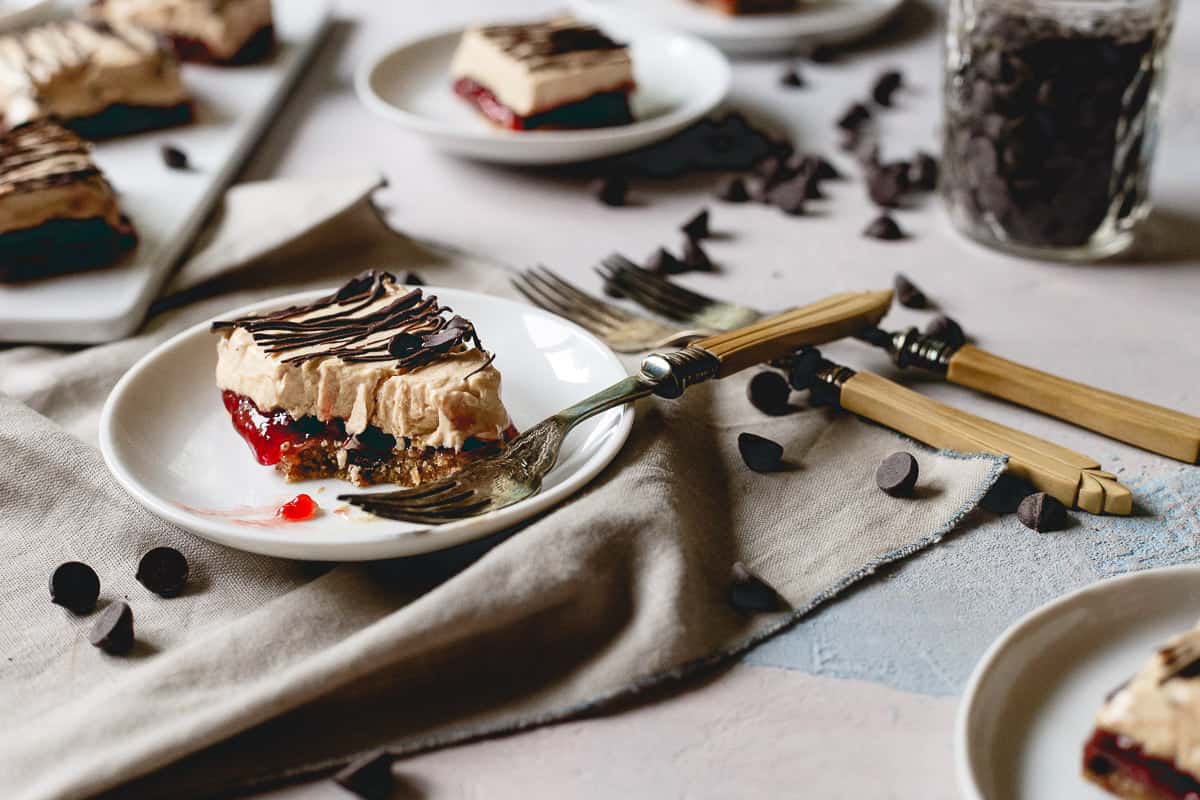 A dessert bar with layers of chocolate, cherry, and cream sits on a white plate, garnished with drizzled chocolate, surrounded by forks and chocolate chips.