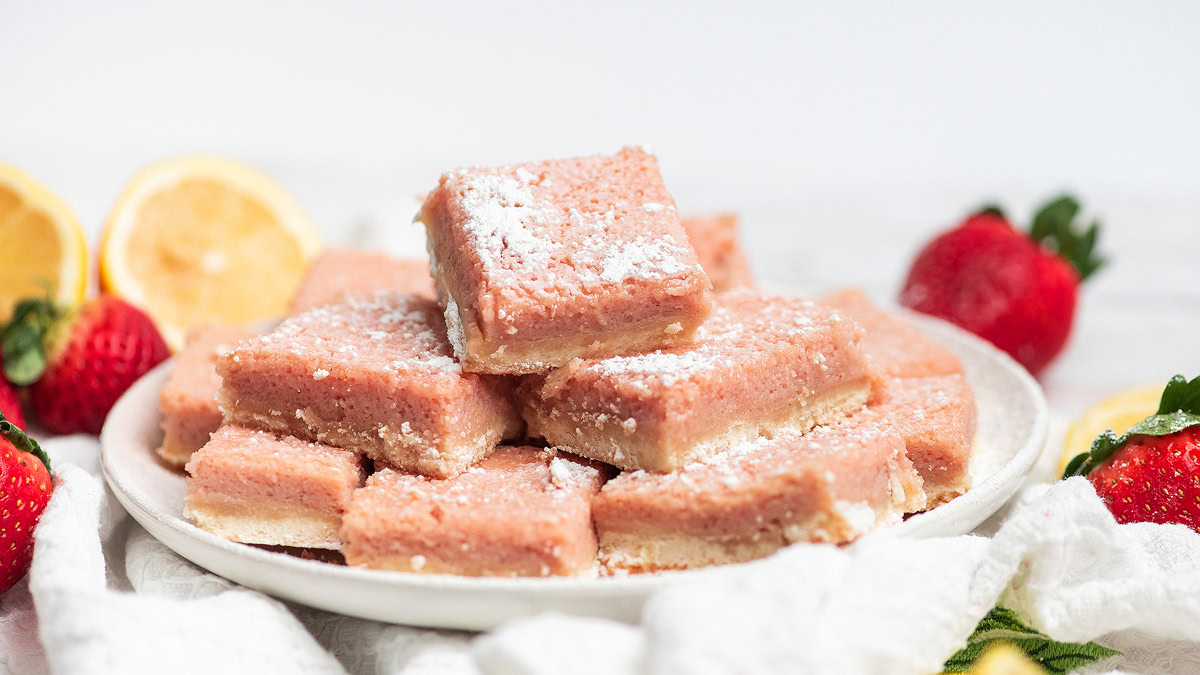 A plate of strawberry lemon bars dusted with powdered sugar, surrounded by fresh strawberries, lemon halves, and a white cloth.