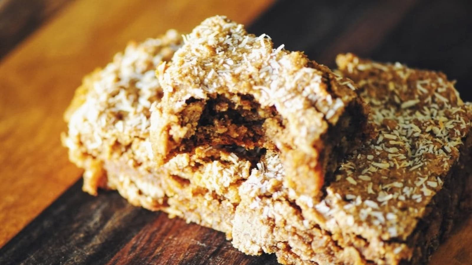 Two pieces of oat and coconut bars on a wooden surface, with one piece showing a bite taken out.