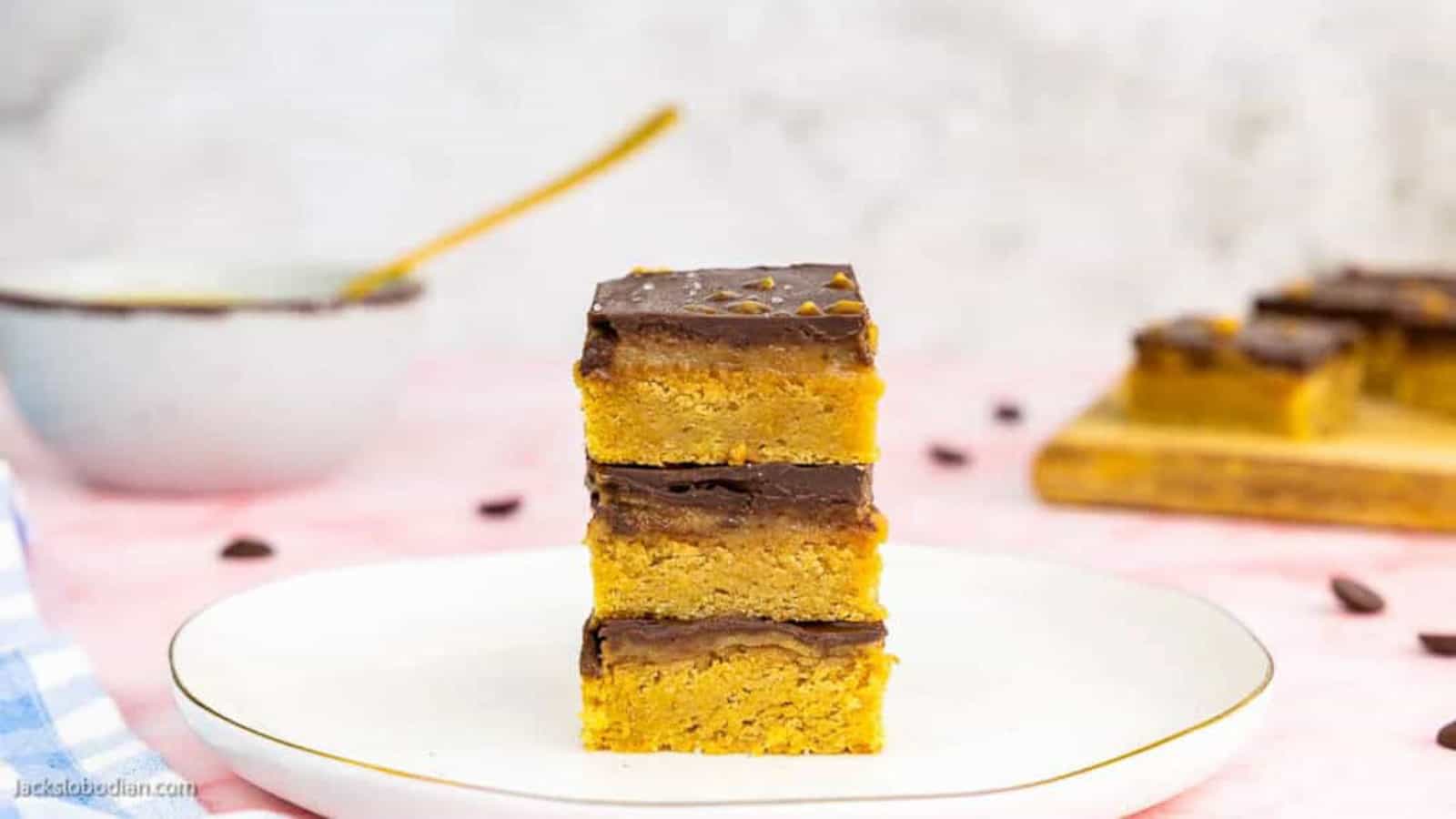 Three stacked bars with a golden base and a chocolate layer on top are placed on a white plate, with more bars and a bowl in the blurred background.