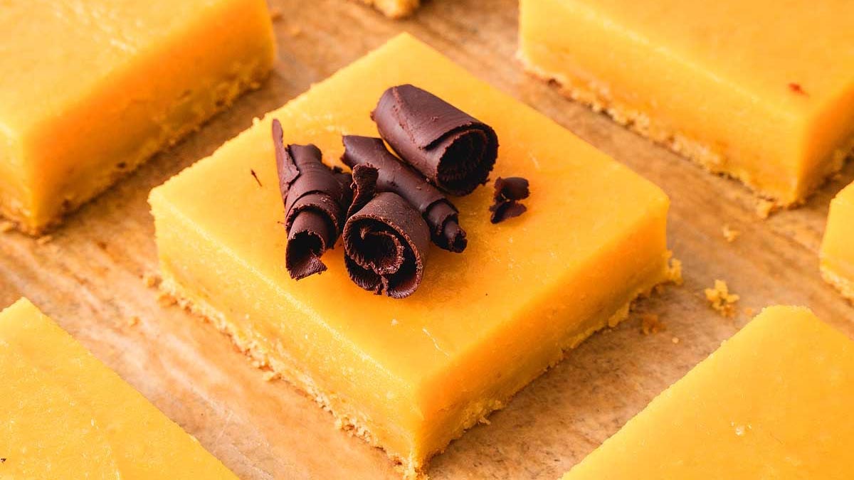 A square lemon bar with a crumbly crust is topped with dark chocolate curls, placed on a parchment-lined surface alongside other lemon bars.