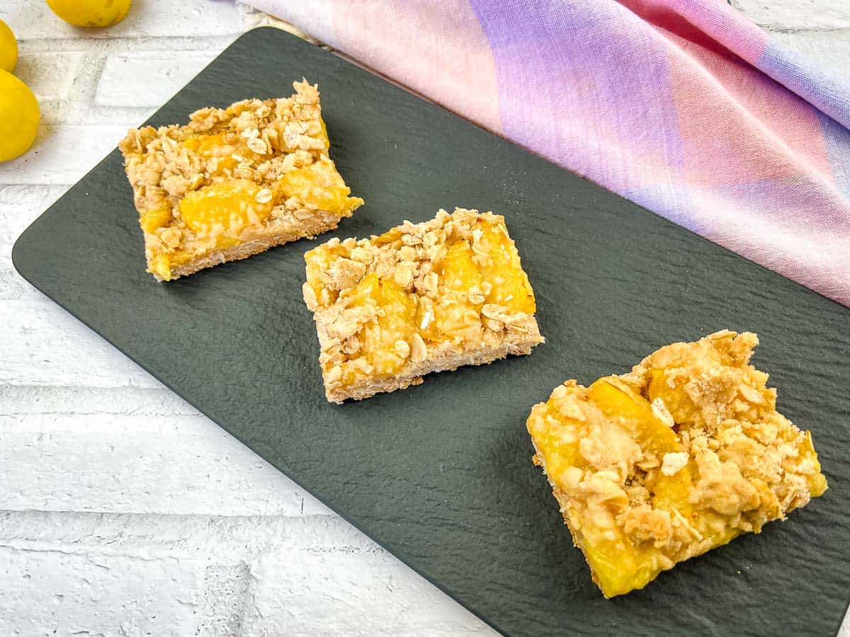 Three rectangular fruit crumble bars are arranged on a dark slate serving board, with a pastel cloth and yellow fruits in the background.