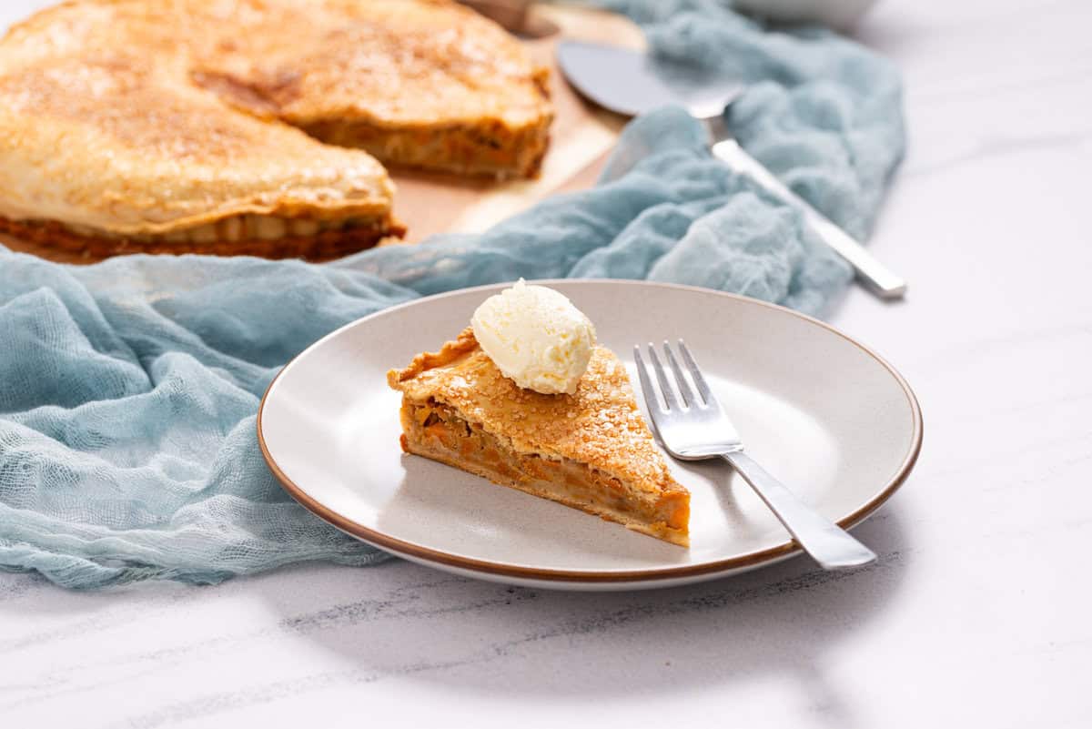 A slice of pie topped with a scoop of vanilla ice cream on a plate with a fork, with the remaining pie in the background.