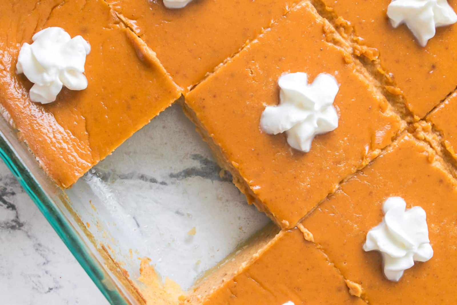 Sliced pumpkin pie bars in a glass dish, each topped with a swirl of whipped cream; one piece is missing.