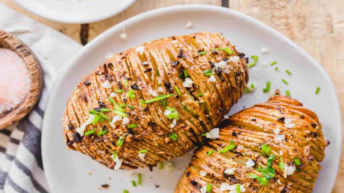 Two hasselback potatoes on a white plate, garnished with chopped chives, minced garlic, and coarse salt.