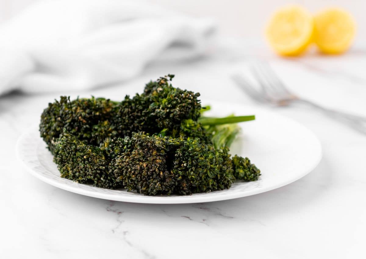 A white plate with roasted broccolini on a marble surface, with a fork and sliced lemons in the blurred background.