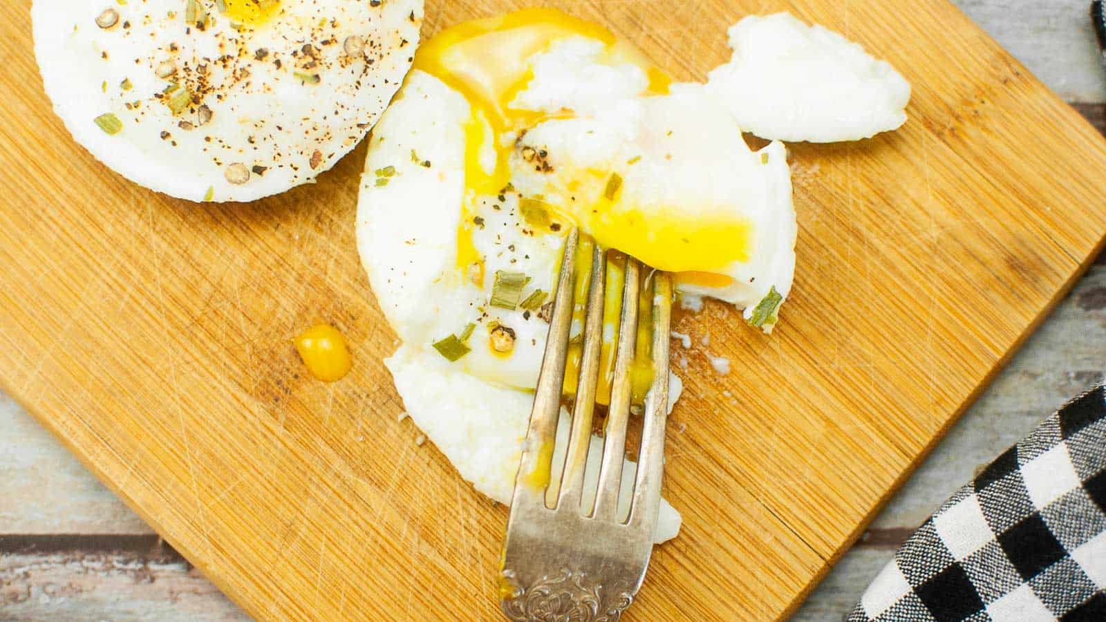 A fork rests on a poached egg with runny yolk and herbs, placed on a wooden cutting board next to a black and white checkered cloth.