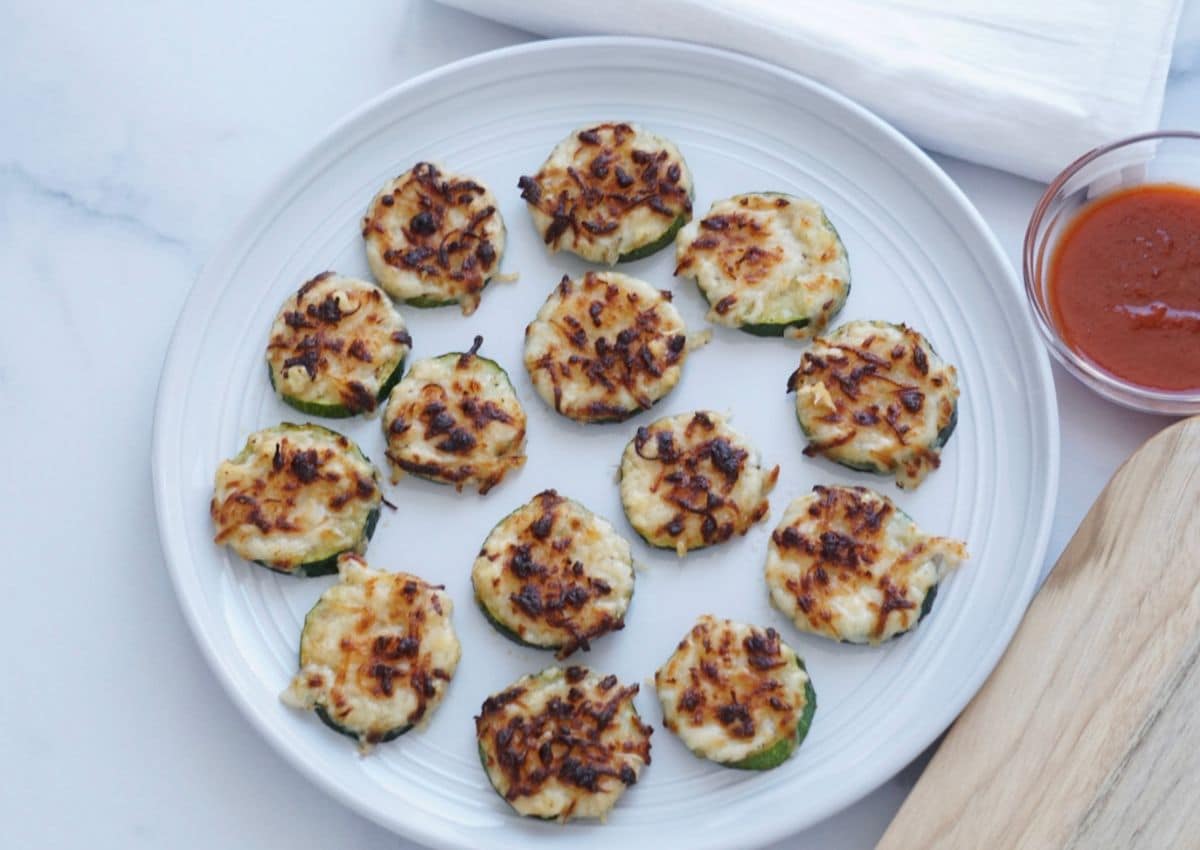 A white plate with baked zucchini rounds topped with melted cheese, arranged in a circle. A small bowl of red dipping sauce is on the side.