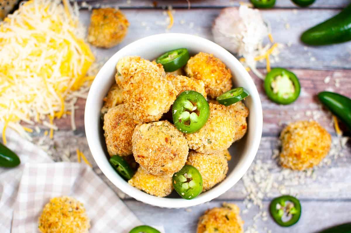 A bowl of breaded, baked cheese balls garnished with sliced fresh jalapeรฑos, surrounded by shredded cheese and jalapeรฑos on a wooden surface.