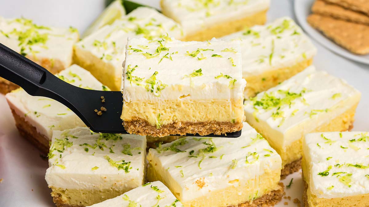 A spatula lifts a square piece of key lime cheesecake bar topped with whipped cream and lime zest, surrounded by similar bars on a tray.