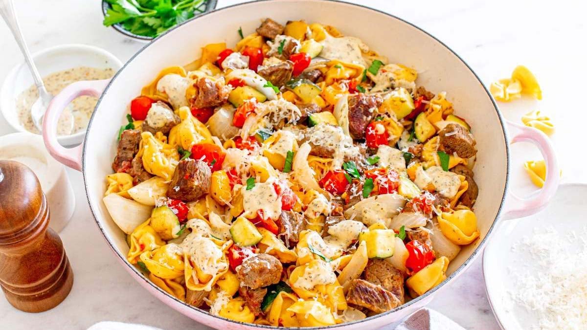 A large pan filled with tortellini pasta, beef cubes, cherry tomatoes, zucchini, and a creamy sauce, garnished with herbs and grated cheese.