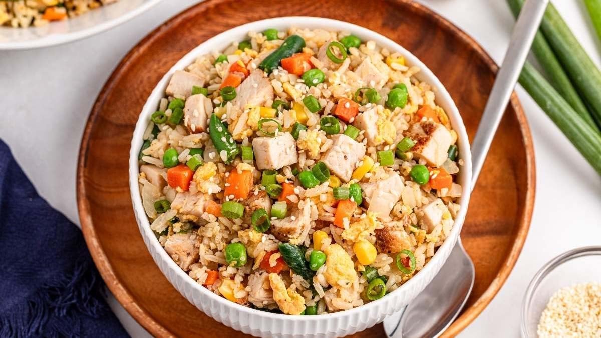 A bowl of chicken fried rice with vegetables including peas, carrots, corn, and green onions, served with a spoon on a wooden plate.