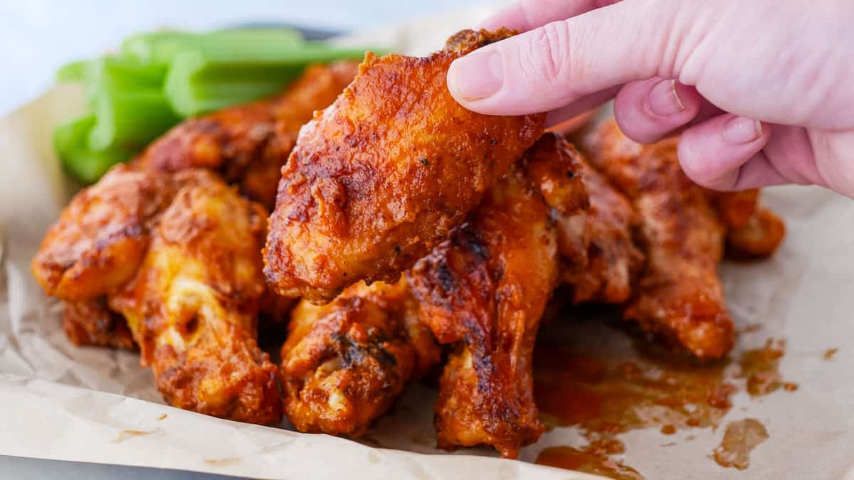 A hand holding a buffalo chicken wing above a pile of sauced chicken wings on parchment paper, with celery sticks in the background.