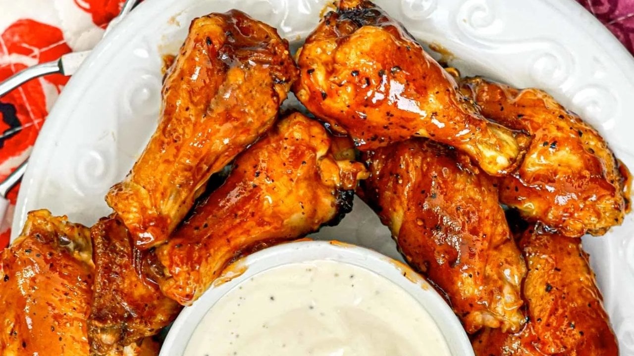 A plate of glazed chicken drumsticks is arranged around a bowl of white dipping sauce on a white dish.