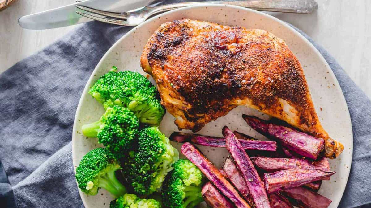A plate with a seasoned baked chicken leg, steamed broccoli, and roasted purple sweet potato fries, with a fork and knife beside the plate.
