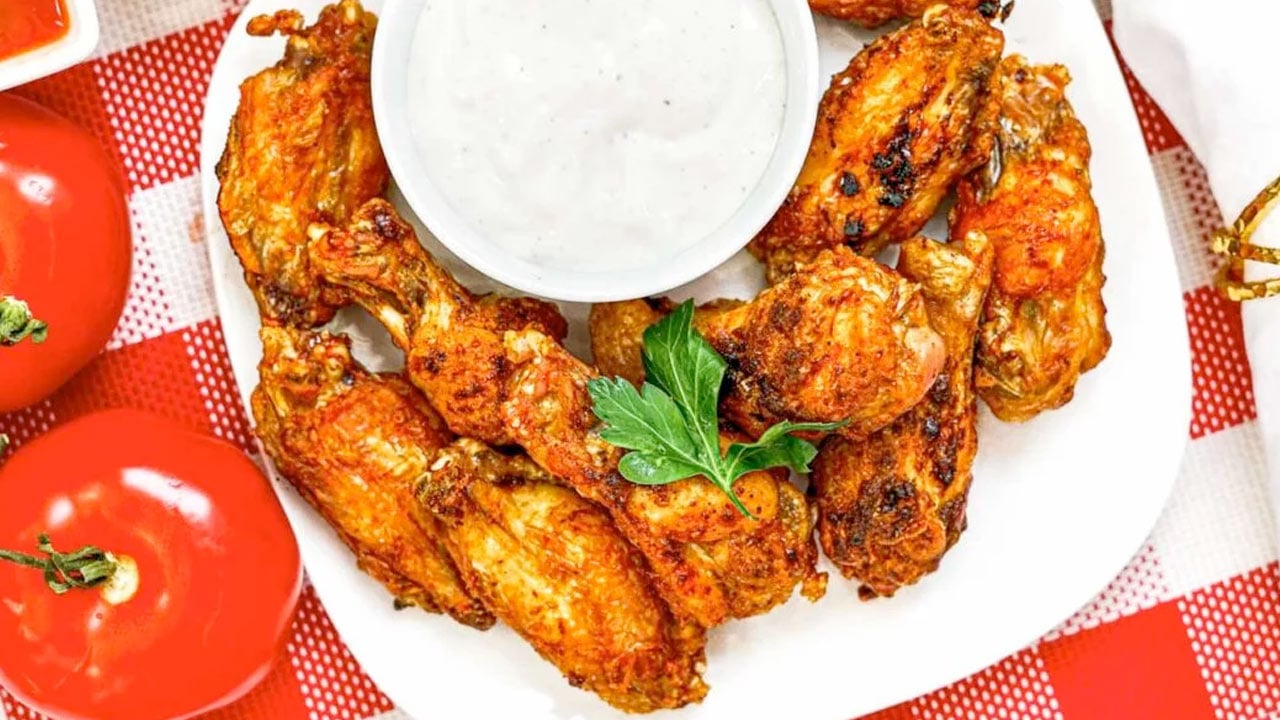 A plate of cooked chicken wings garnished with parsley, served with a cup of white dipping sauce, placed on a red and white checkered tablecloth next to fresh tomatoes.