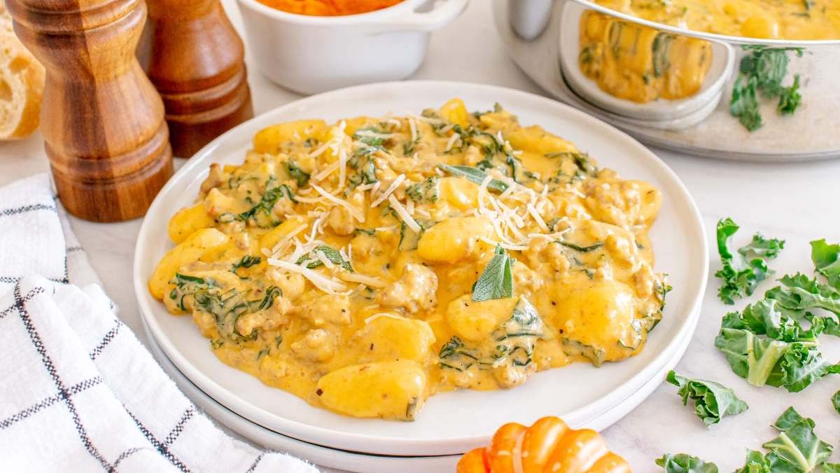 A plate of creamy gnocchi with wilted greens and grated cheese, surrounded by fresh kale, a pot of sauce, and pepper mills on a white countertop.