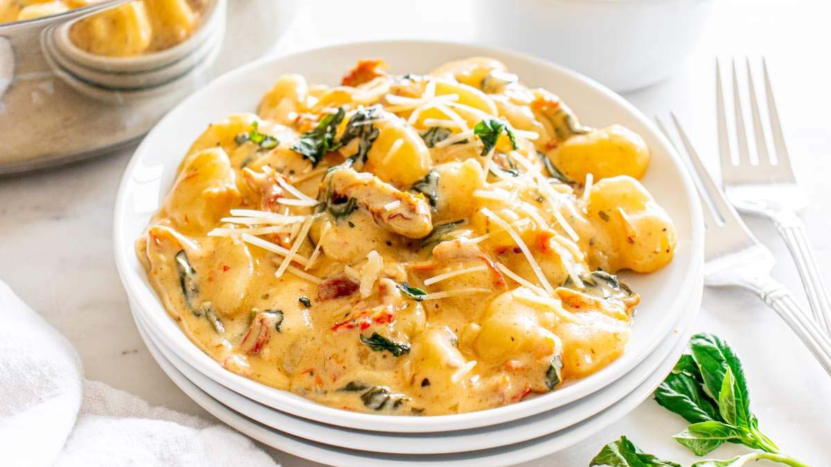 A white plate of creamy gnocchi topped with grated cheese and herbs sits on a table next to a fork and fresh basil leaves.