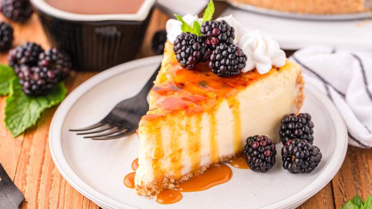 A slice of cheesecake topped with caramel sauce, blackberries, and whipped cream sits on a plate with a fork.
