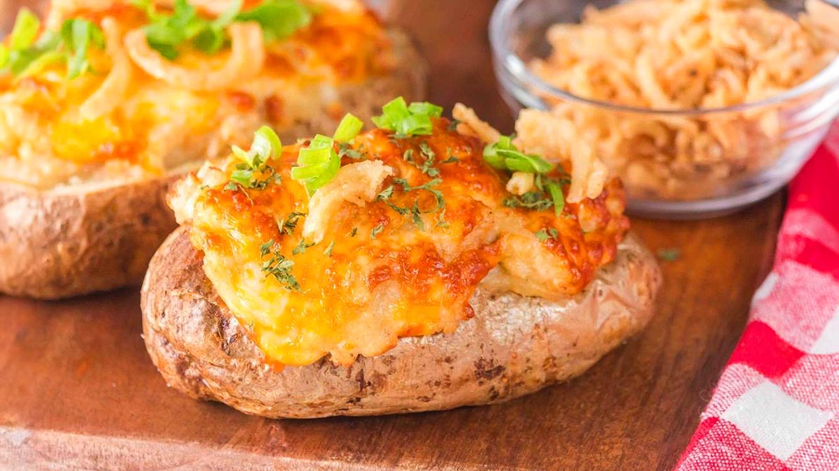 Two baked potatoes topped with melted cheese, green onions, and crispy fried onions on a wooden surface, with a bowl of fried onions in the background.