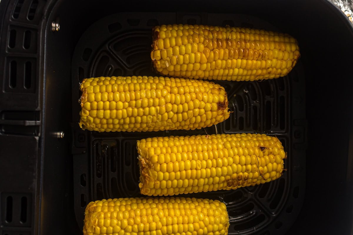 Four cooked corn cobs are arranged in a single layer inside an air fryer basket.