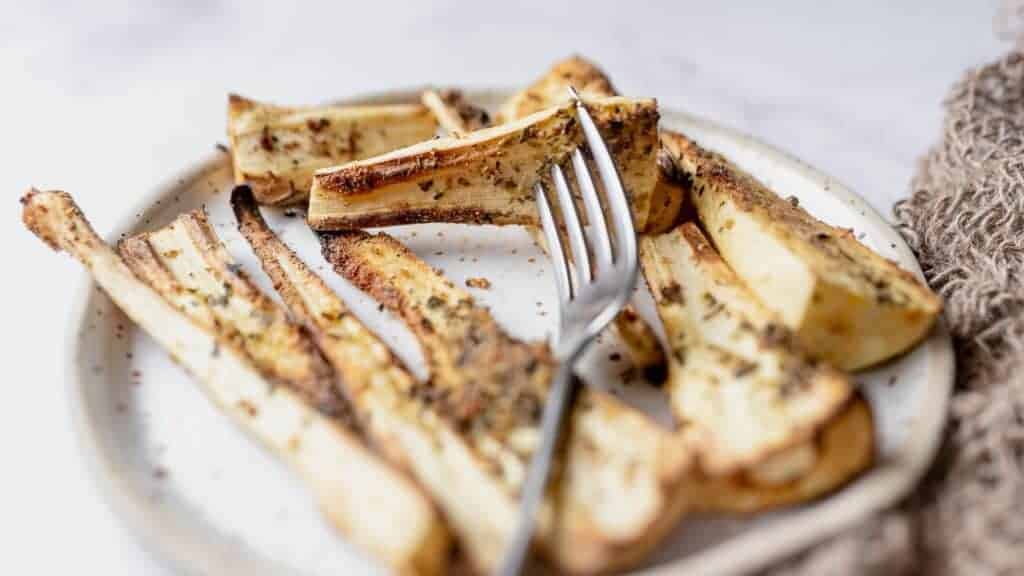A plate of roasted parsnip wedges seasoned with herbs and spices, with a fork resting among the pieces.