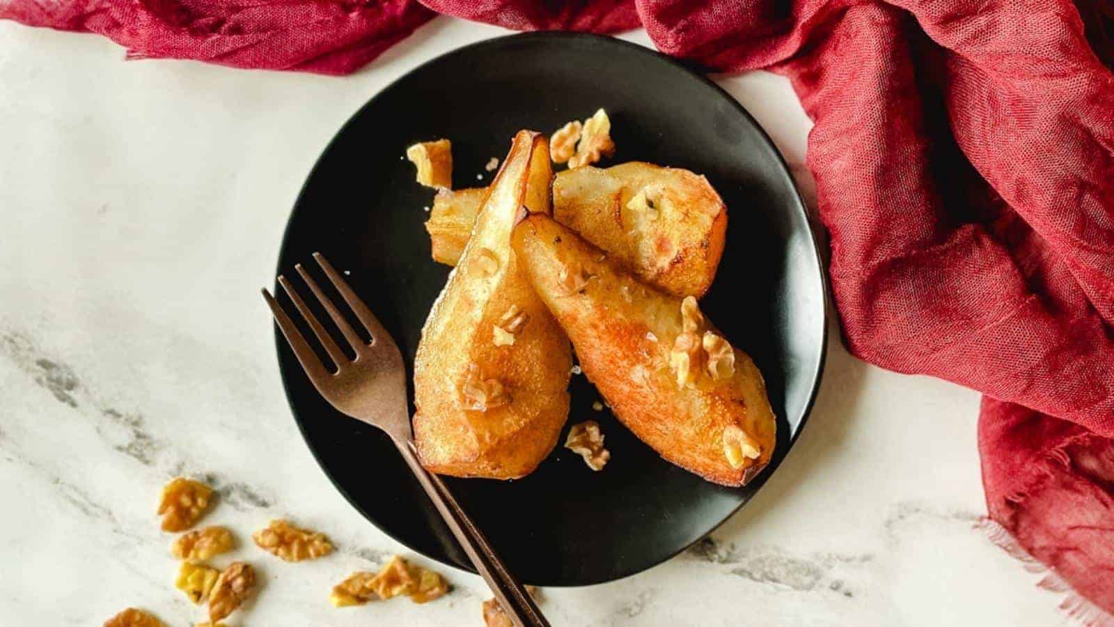 A black plate with roasted pear halves topped with chopped walnuts, a fork beside them, and a red cloth in the background.