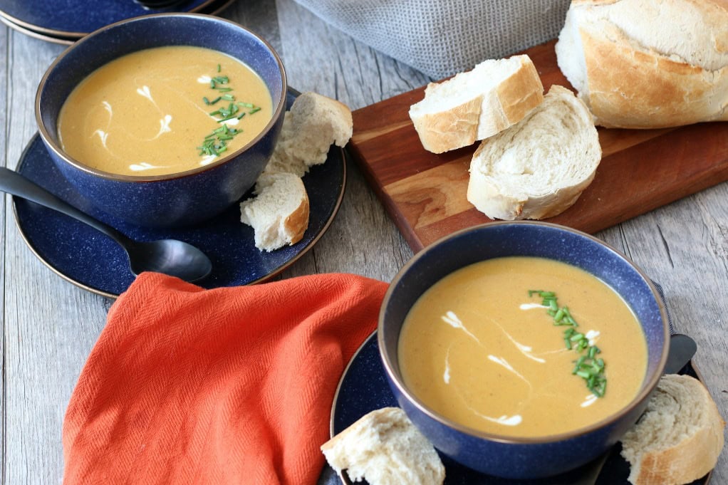 Two bowls of creamy soup garnished with herbs, served with slices of bread on blue plates, with an orange napkin and a wooden board on a rustic table.