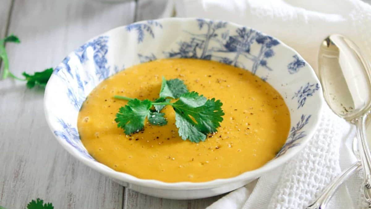 A bowl of creamy orange soup garnished with cilantro leaves and ground black pepper, placed on a white table with a spoon beside it.