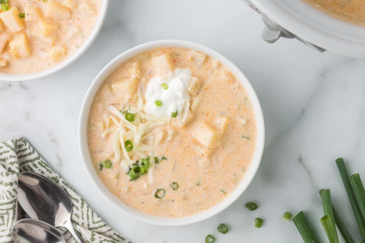 A bowl of creamy potato soup topped with shredded cheese, sour cream, and chopped green onions on a white marble surface with spoons and green onions nearby.