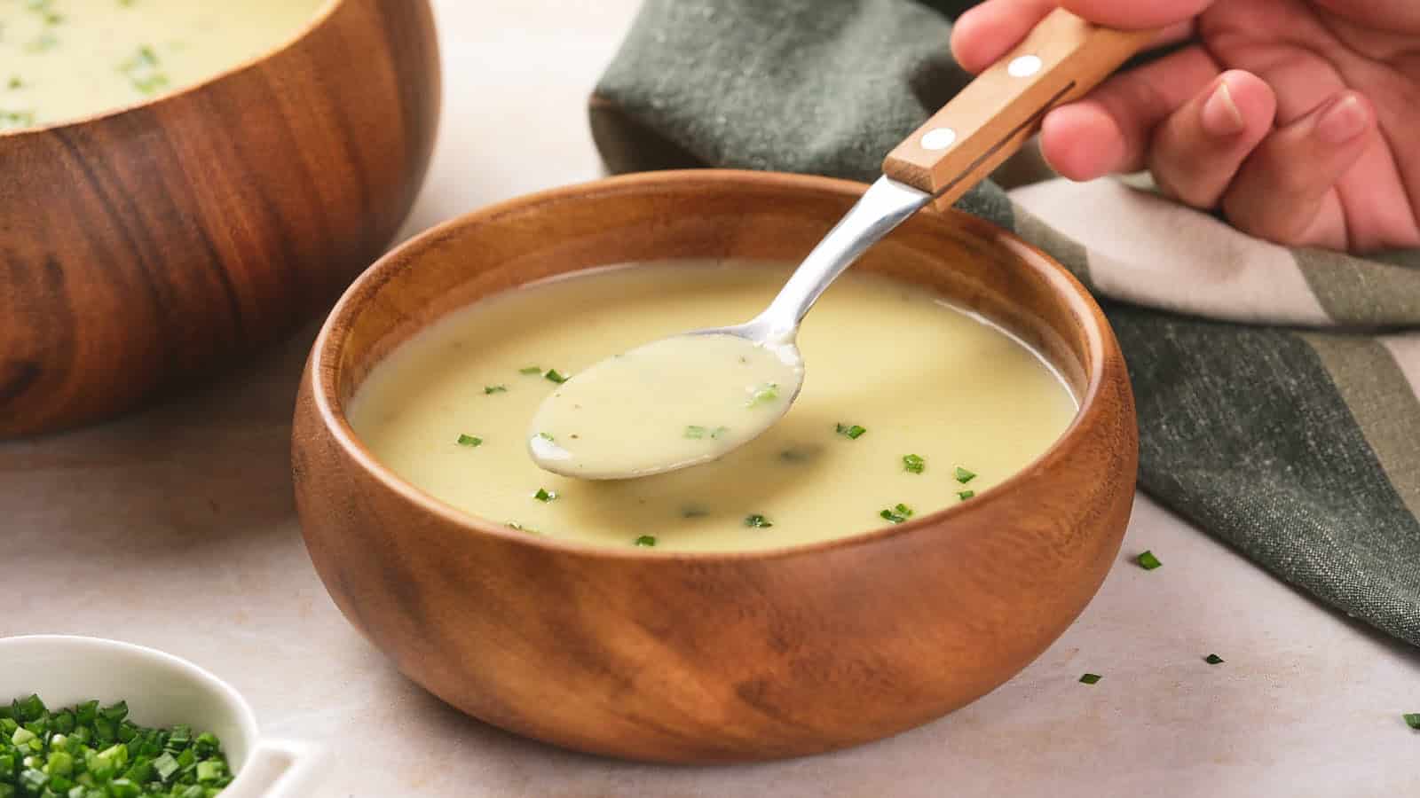 A hand holds a spoonful of creamy soup garnished with chopped chives, served in a wooden bowl on a light surface.