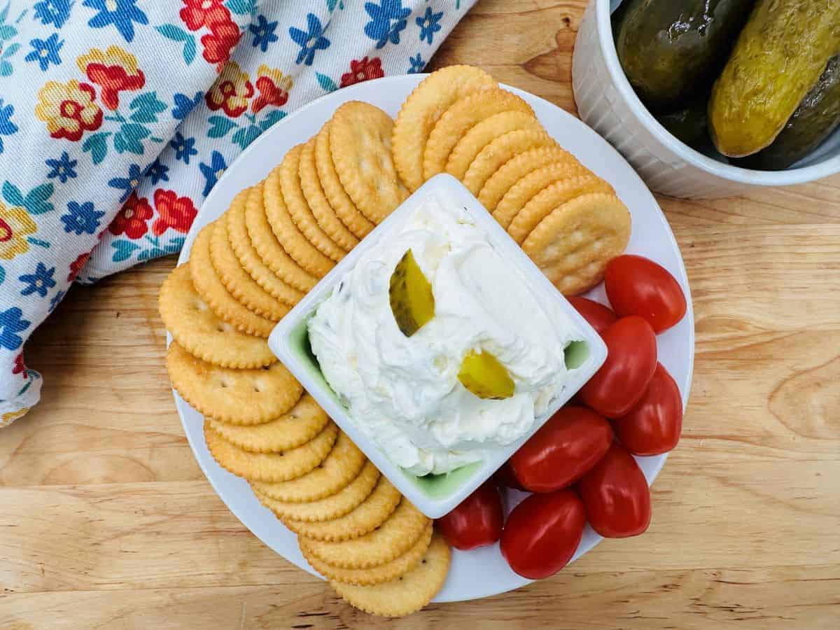 A plate with round crackers, grape tomatoes, and a bowl of creamy dip garnished with pickle slices, next to a bowl of pickles on a wooden surface.