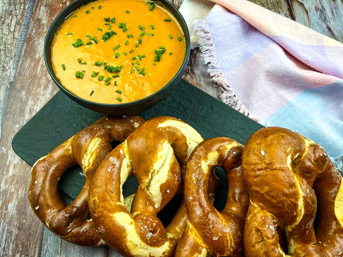 A bowl of orange cheese dip garnished with chives sits beside three soft pretzels on a black serving board, with a colorful cloth in the background.