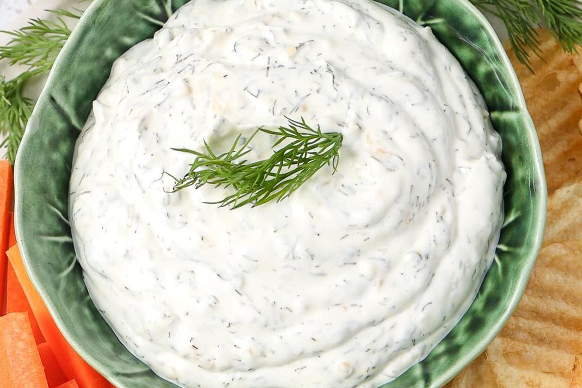 A green bowl filled with creamy dip, garnished with dill, surrounded by potato chips and carrot sticks.
