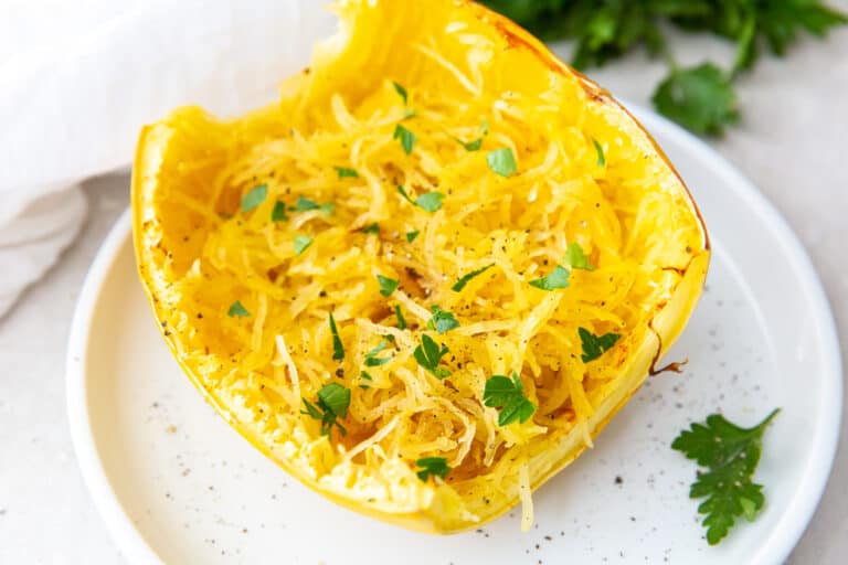 Half of a cooked spaghetti squash on a white plate, topped with chopped parsley and black pepper.
